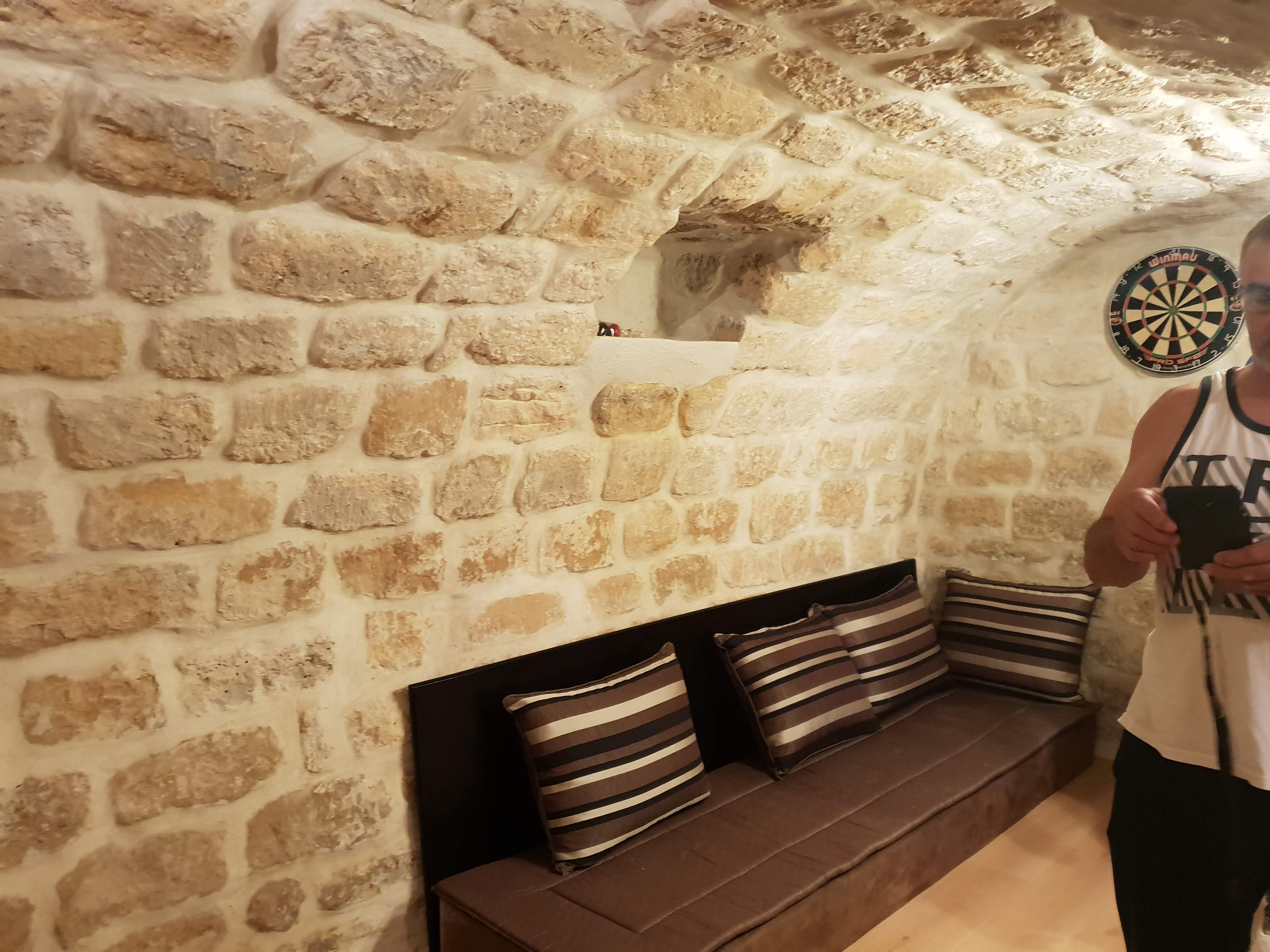 Cave aux Lilas devenue un salon avec banquette et coussins, mur en pierre apparente rejointoyé